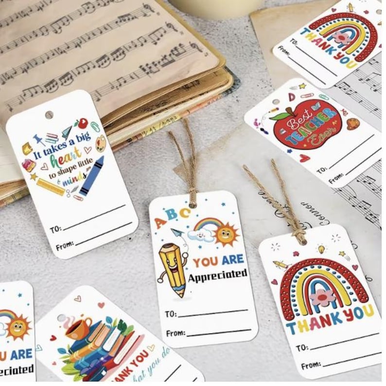 Peut inclure: Une collection d'&eacute;tiquettes cadeaux rectangulaires blanches orn&eacute;es d'illustrations color&eacute;es sur le th&egrave;me de l'&eacute;cole, incluant des arcs-en-ciel, des pommes, des crayons et des livres. Chaque &eacute;tiquette poss&egrave;de un trou avec de la ficelle, du texte imprim&eacute; comme "Thank You" ou "Best Teacher Ever" et des lignes pour "To" et "From".