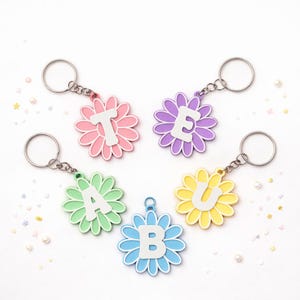 May include: Five flower-shaped keyrings in various colours: pink, purple, green, blue, and yellow. Each keyring features a different letter: T, E, A, B, and U, in white. The keyrings have silver metal attachments.