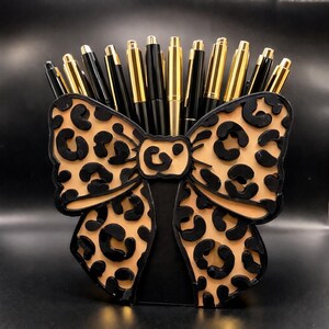 May include: A pen holder shaped like a bow, featuring a tan leopard print design with black accents. The holder is filled with gold and black pens. The bow-shaped holder is made of wood or a similar material.