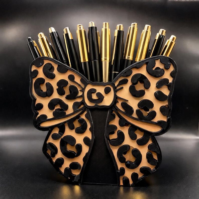 May include: A pen holder shaped like a bow, featuring a tan leopard print design with black accents. The holder is filled with gold and black pens. The bow-shaped holder is made of wood or a similar material.