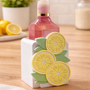 May include: A pink liquid soap dispenser with a white pump sits in a white holder decorated with yellow lemon slices and green leaves. The dispenser has a gold band. Lemons are visible in the background.