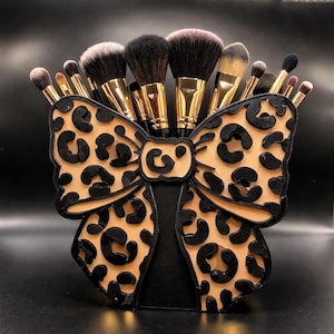 May include: A black and tan leopard print makeup brush holder shaped like a bow, filled with various makeup brushes. The holder has a black base and a tan bow with black leopard spots. The brushes have gold handles and black and tan bristles.