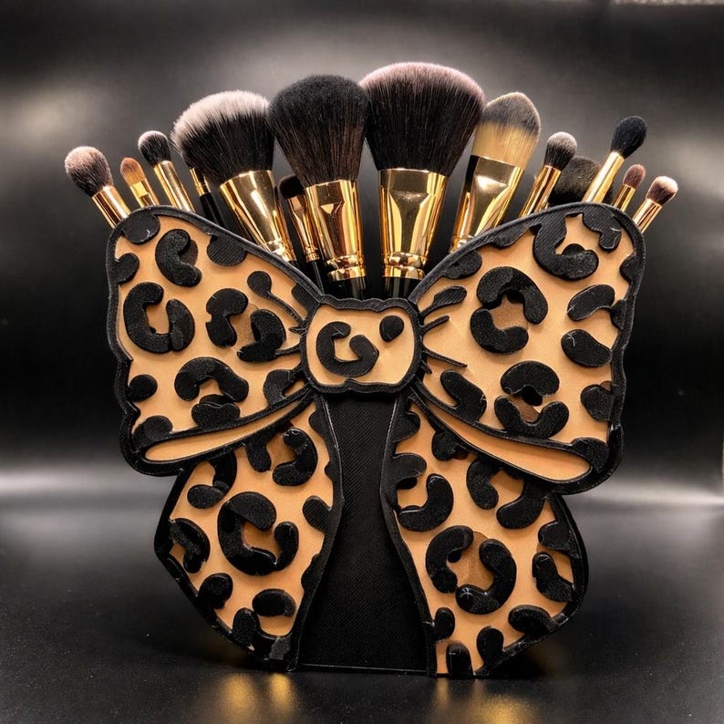 May include: A black and tan leopard print makeup brush holder shaped like a bow, filled with various makeup brushes. The holder has a black base and a tan bow with black leopard spots. The brushes have gold handles and black and tan bristles.