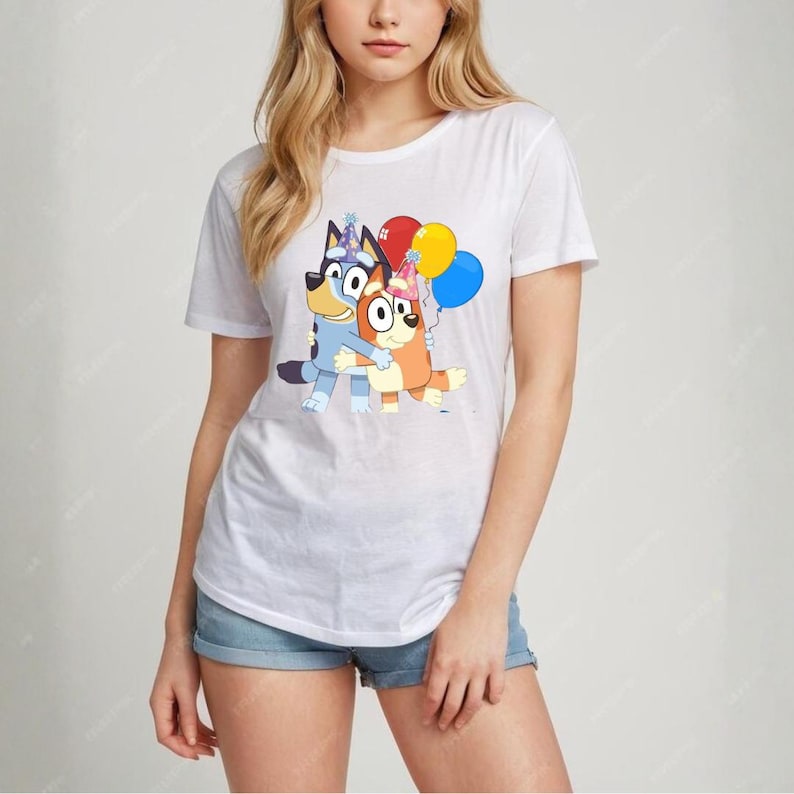 May include: White t-shirt featuring a cartoon graphic of two cartoon dogs hugging, with party hats and balloons. The dogs are blue and orange. The balloons are red, yellow, and blue. The shirt is a crew neck.
