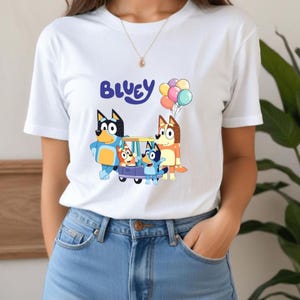 May include: White t-shirt featuring the word "Bluey" in blue script above a colorful cartoon image of Bluey characters in a car with balloons. The design includes characters in various colors.