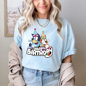 May include: Light blue t-shirt featuring a colorful graphic of two cartoon dogs wearing party hats, with the words "Happy Birthday" in a playful font. The shirt is made of soft material.