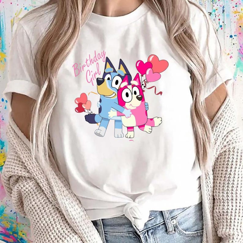 May include: White t-shirt featuring a cartoon graphic of two hugging cartoon dogs, one blue and one pink, holding heart-shaped balloons. The text "Birthday Girl" is above the dogs in pink script. The shirt is made of a soft material.