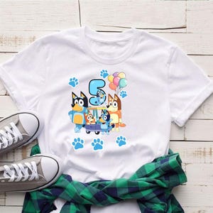 May include: White t-shirt featuring a colorful graphic of cartoon dogs, the number 5, balloons, and paw prints. The design includes a blue car with the cartoon dogs. The shirt is paired with gray sneakers and a green plaid shirt.