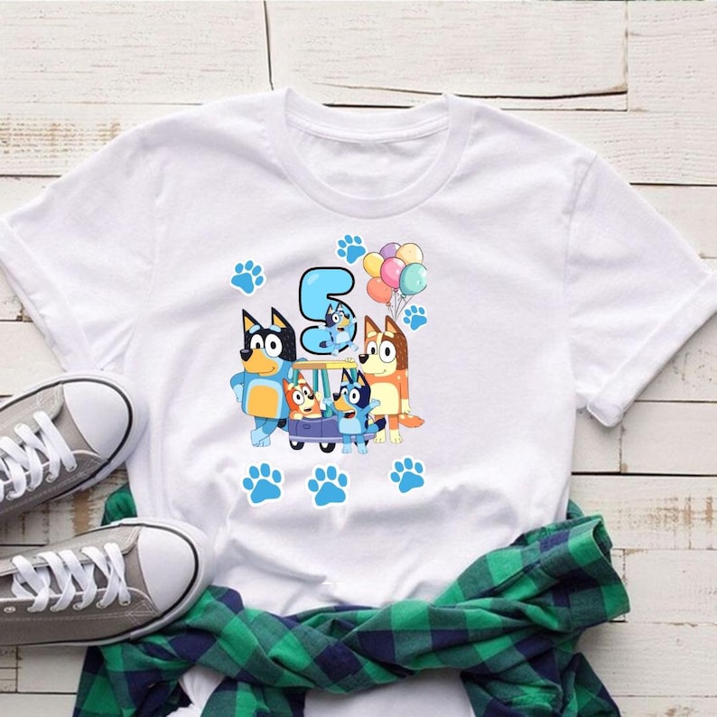 May include: White t-shirt featuring a colorful graphic of cartoon dogs, the number 5, balloons, and paw prints. The design includes a blue car with the cartoon dogs. The shirt is paired with gray sneakers and a green plaid shirt.