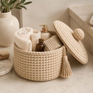 May include: A round, beige crocheted storage basket with a matching lid featuring a central knob and a side tassel. The basket contains rolled cream-colored towels, a small amber glass bottle, a pump dispenser, and a textured square cloth.
