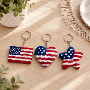 May include: Three crochet keychains featuring the American flag pattern in red, white, and blue. The set includes a rectangular flag, a heart-shaped piece, and a star-shaped piece, each attached to a silver-toned metal split ring keychain.