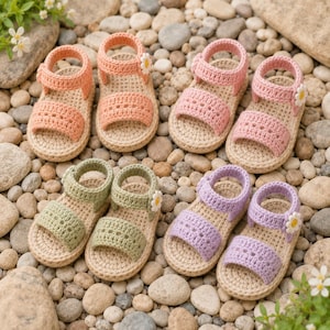 May include: Four pairs of handmade crochet sandals in peach, pink, sage green, and lavender. Each sandal features a beige sole, a two-strap design, and a small white daisy flower accent on the side of the ankle strap, arranged on a stone surface.
