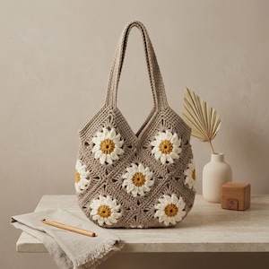 Op de afbeelding: Een gehaakte draagtas in beige, versierd met witte en gele madeliefmotieven. De tas heeft twee handvatten en wordt getoond op een licht oppervlak. Een haaknaald en een linnen doek zijn ook zichtbaar.
