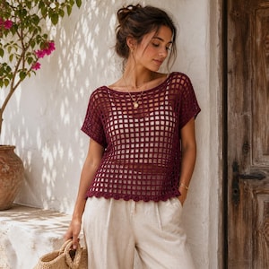 Può includere: Un top all'uncinetto bordeaux con un motivo a griglia quadrata. Il top ha maniche corte e un orlo smerlato. La modella indossa il top con pantaloni beige e una borsa di paglia. Lo sfondo comprende un muro bianco e una porta di legno.