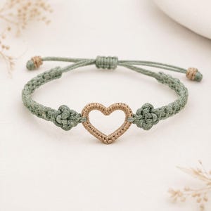 May include: A macrame bracelet featuring a central heart-shaped charm in a light tan color. The band is constructed from sage green cord with intricate knotting details on either side of the heart and adjustable sliding knots at the ends.