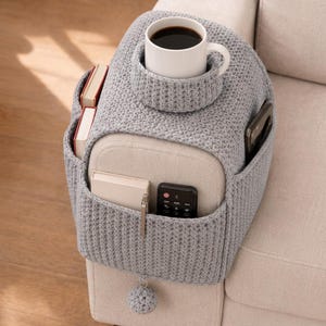 May include: A gray knitted sofa arm organizer with a built-in cup holder holding a white mug of coffee. It has pockets for books, a remote, a phone, and a pen. A small gray pom-pom hangs from the bottom.