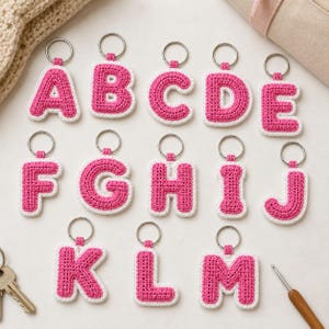 May include: A collection of thirteen individual crochet letter keychains, each featuring a vibrant pink yarn center with a white yarn border. Every letter from A through M is attached to a silver-toned metal split ring for easy attachment.