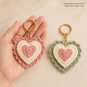 May include: Two heart-shaped crochet keychains with gold-tone metal rings. One keychain features layers of dusty pink and cream yarn, while the other has layers of sage green, cream, and dusty pink yarn. Text reads: Al-generated model shown Digital PDF Pattern Only.
