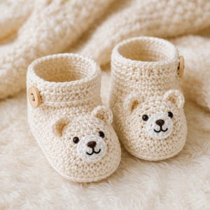 May include: A pair of cream-coloured crochet booties featuring a bear face design on the front. Each bootie has two small rounded ears, embroidered black eyes, a brown nose, and a stitched mouth. A single light brown wooden button is attached to the side.