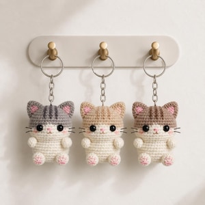 May include: Three handmade crochet cat keychains in gray, tan, and brown, each with pink accents on the ears and paws. The keychains are attached to silver keyrings and hang from a wall-mounted hook rack with gold-colored knobs. The cats have black embroidered whiskers.