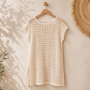 May include: A cream-colored, short-sleeved tunic featuring an open-knit, grid-like crochet pattern. The garment has a round neckline, a straight hemline, and vertical side slits at the bottom, hanging on a wooden hanger against a textured wall.