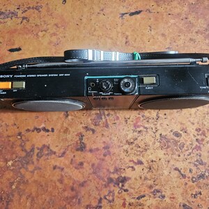 Puede incluir: Sistema de altavoces est&eacute;reo Sony negro con asa y antena retr&aacute;ctil. El est&eacute;reo tiene dos altavoces, un bot&oacute;n de ajuste del temporizador y un control de volumen. El texto "SONY Powered Stereo Speaker System SRF-80W" est&aacute; impreso.