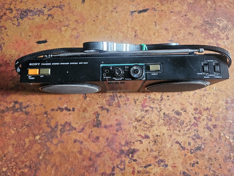 Puede incluir: Sistema de altavoces est&eacute;reo Sony negro con asa y antena retr&aacute;ctil. El est&eacute;reo tiene dos altavoces, un bot&oacute;n de ajuste del temporizador y un control de volumen. El texto "SONY Powered Stereo Speaker System SRF-80W" est&aacute; impreso.