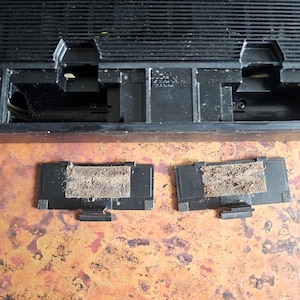 Puede incluir: Un dispositivo electr&oacute;nico negro de forma rectangular, con una rejilla superior y dos compartimentos abiertos. Dos peque&ntilde;as cubiertas negras rectangulares con un filtro beige polvoriento est&aacute;n delante del dispositivo.