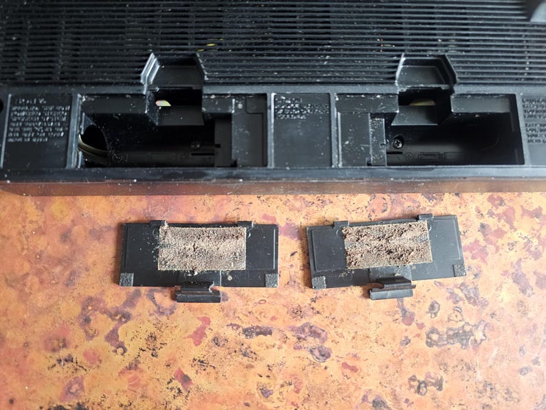 Puede incluir: Un dispositivo electr&oacute;nico negro de forma rectangular, con una rejilla superior y dos compartimentos abiertos. Dos peque&ntilde;as cubiertas negras rectangulares con un filtro beige polvoriento est&aacute;n delante del dispositivo.