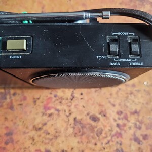 Puede incluir: Primer plano de un radiocasete negro vintage con antena plateada y controles. Incluye un bot&oacute;n "EJECT", controles de tono y agudos, y una rejilla de altavoz. El radiocasete est&aacute; sobre una superficie marr&oacute;n texturizada.