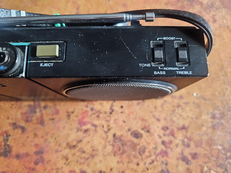 Puede incluir: Primer plano de un radiocasete negro vintage con antena plateada y controles. Incluye un bot&oacute;n "EJECT", controles de tono y agudos, y una rejilla de altavoz. El radiocasete est&aacute; sobre una superficie marr&oacute;n texturizada.