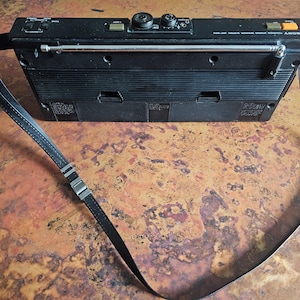 Puede incluir: Radio Sony vintage negra con correa para el hombro. La radio tiene una antena telesc&oacute;pica, perillas de control y botones. La correa es negra con herrajes met&aacute;licos de color plateado. La radio es rectangular con un dise&ntilde;o acanalado en la parte delantera.