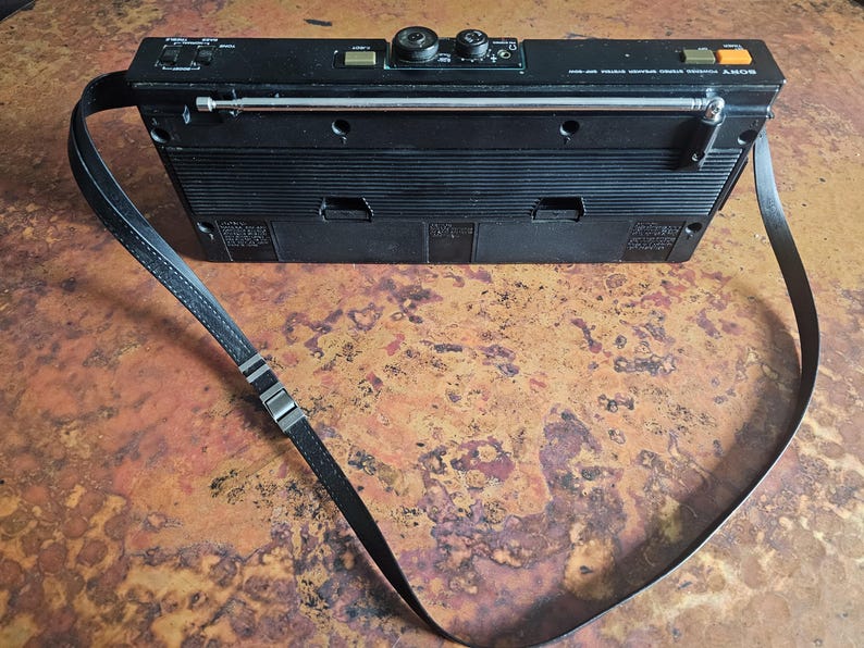 Puede incluir: Radio Sony vintage negra con correa para el hombro. La radio tiene una antena telesc&oacute;pica, perillas de control y botones. La correa es negra con herrajes met&aacute;licos de color plateado. La radio es rectangular con un dise&ntilde;o acanalado en la parte delantera.