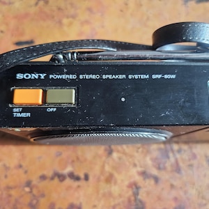 Puede incluir: Sistema de altavoces est&eacute;reo con alimentaci&oacute;n Sony SRF-80W negro con correa de cuero. Cuenta con botones naranja y verde etiquetados "SET TIMER" y "OFF". El texto "SONY POWERED STEREO SPEAKER SYSTEM SRF-80W" est&aacute; impreso.