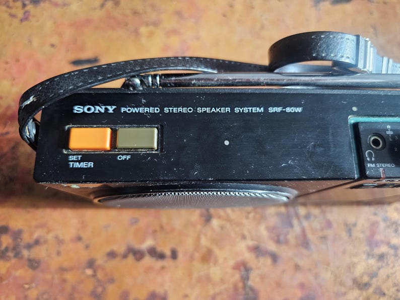 Puede incluir: Sistema de altavoces est&eacute;reo con alimentaci&oacute;n Sony SRF-80W negro con correa de cuero. Cuenta con botones naranja y verde etiquetados "SET TIMER" y "OFF". El texto "SONY POWERED STEREO SPEAKER SYSTEM SRF-80W" est&aacute; impreso.