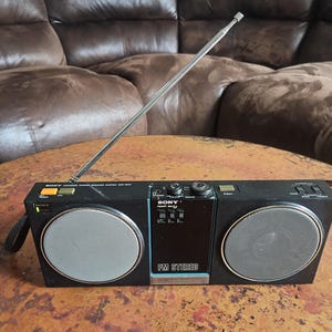 May include: Black Sony FM stereo radio with a silver antenna and two circular speakers. The radio has a handle and various buttons and dials. The radio is sitting on a brown table.