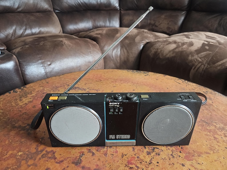 Puede incluir: Radio est&eacute;reo FM Sony negra con antena plateada y dos altavoces circulares. La radio tiene un asa y varios botones y diales. La radio est&aacute; sobre una mesa marr&oacute;n.