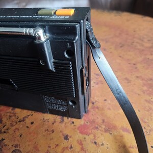 Puede incluir: Grabadora de casetes de radio est&eacute;reo Sony negra vintage con antena plateada y correa negra. La radio tiene botones verdes y naranjas y una antena plateada. La radio est&aacute; sobre una superficie marr&oacute;n.