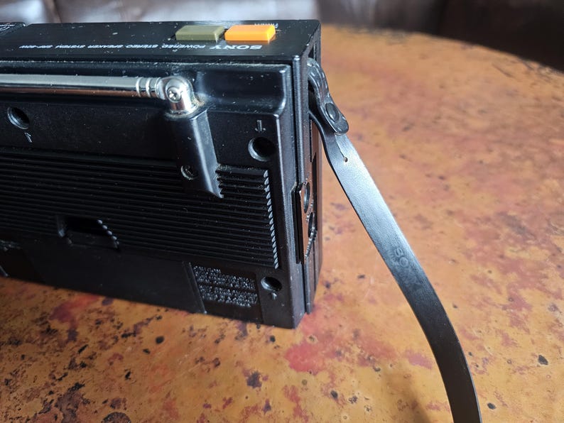 Puede incluir: Grabadora de casetes de radio est&eacute;reo Sony negra vintage con antena plateada y correa negra. La radio tiene botones verdes y naranjas y una antena plateada. La radio est&aacute; sobre una superficie marr&oacute;n.
