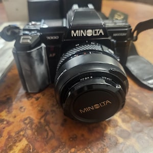 May include: Black Minolta 7000 AF camera with a lens cap. The camera has the Minolta logo in white. The lens has a 49 mm diameter. The camera strap is black. The camera is on a brown surface.