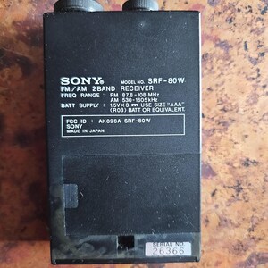 Puede incluir: Receptor FM/AM de 2 bandas Sony SRF-80W negro. El dispositivo tiene dos perillas negras en la parte superior y una etiqueta con el nombre de la marca, el n&uacute;mero de modelo y el rango de frecuencia. Tambi&eacute;n es visible el texto "MADE IN JAPAN".