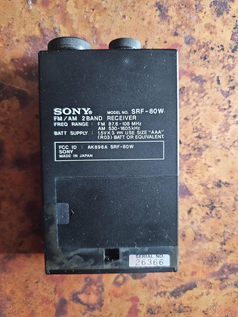 Puede incluir: Receptor FM/AM de 2 bandas Sony SRF-80W negro. El dispositivo tiene dos perillas negras en la parte superior y una etiqueta con el nombre de la marca, el n&uacute;mero de modelo y el rango de frecuencia. Tambi&eacute;n es visible el texto "MADE IN JAPAN".