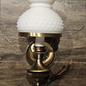 May include: A wall-mounted lamp featuring a white milk glass shade with a hobnail texture and a ruffled top edge. The fixture has a brass-toned metal base and arm, with a brown electrical cord extending from the bottom for plug-in use.