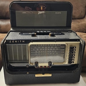 May include: A vintage Zenith Trans-Oceanic portable radio with a black textured case, a gold-toned frequency dial, and a top-hinged lid featuring a chart panel. The front displays the Zenith logo, a speaker grille, and multiple black push-button controls.