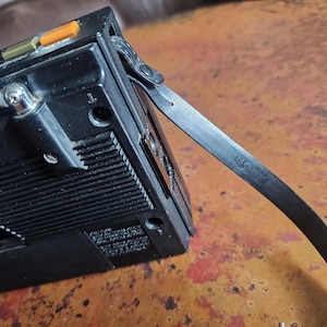 Puede incluir: Radio port&aacute;til vintage negra con antena extensible y correa de cuero. La radio tiene botones naranjas y verdes y un dial plateado. La correa tiene la palabra "SONY" impresa.