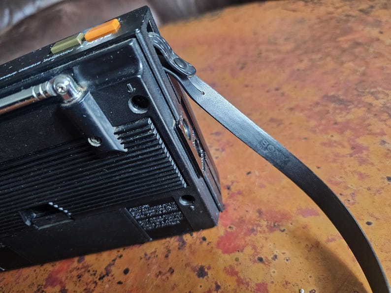 Puede incluir: Radio port&aacute;til vintage negra con antena extensible y correa de cuero. La radio tiene botones naranjas y verdes y un dial plateado. La correa tiene la palabra "SONY" impresa.