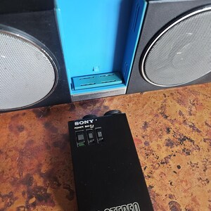 Puede incluir: Una radio est&eacute;reo FM Sony negra y plateada vintage con un compartimento interior azul. La radio tiene dos altavoces con cubiertas de malla plateada. Un mando a distancia negro con el texto "SONY POWER BAND" y "FM STEREO" est&aacute; delante de la radio.