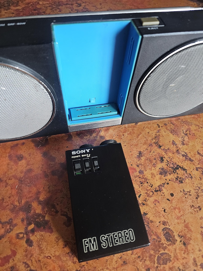 Puede incluir: Una radio est&eacute;reo FM Sony negra y plateada vintage con un compartimento interior azul. La radio tiene dos altavoces con cubiertas de malla plateada. Un mando a distancia negro con el texto "SONY POWER BAND" y "FM STEREO" est&aacute; delante de la radio.