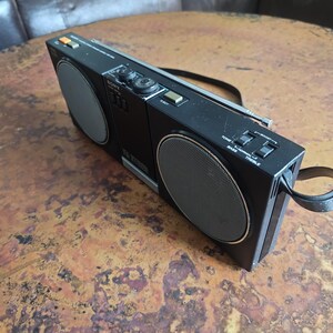 Puede incluir: Boombox Sony negro vintage con dos altavoces y un asa. El boombox tiene detalles plateados, diales y botones. Los altavoces tienen un dise&ntilde;o circular con una cubierta de malla. El boombox est&aacute; sobre una superficie marr&oacute;n.