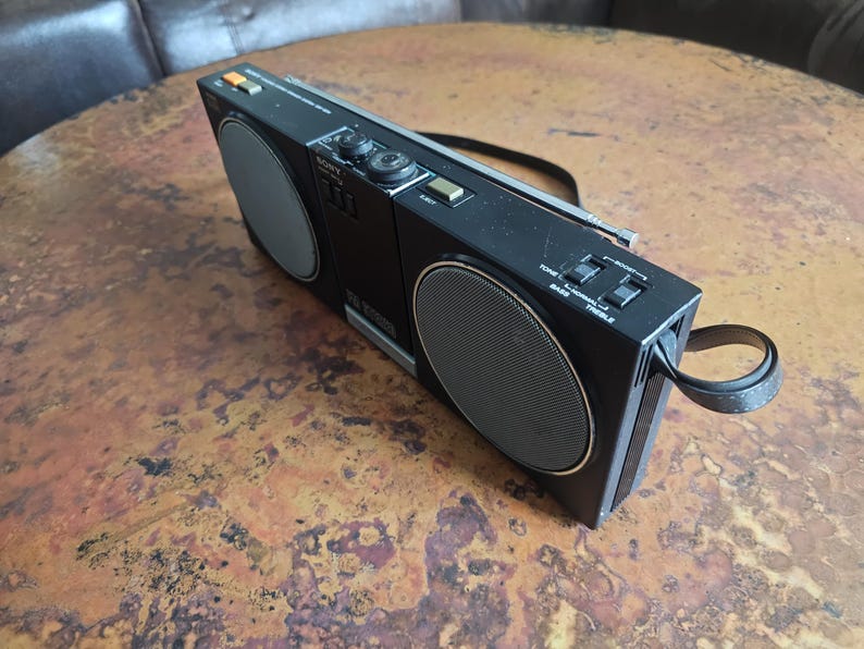 Puede incluir: Boombox Sony negro vintage con dos altavoces y un asa. El boombox tiene detalles plateados, diales y botones. Los altavoces tienen un dise&ntilde;o circular con una cubierta de malla. El boombox est&aacute; sobre una superficie marr&oacute;n.